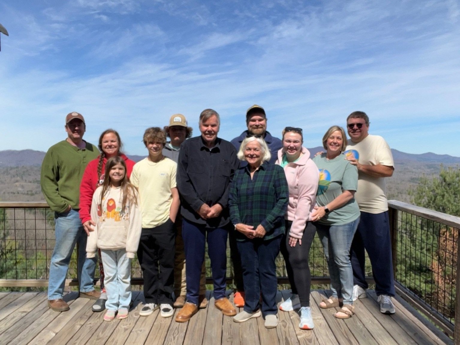 Krista+Fuller+-+IMG_6748 A family gathering in the mountains, featuring nine individuals of various ages posing on a wooden deck with a scenic view of rolling hills and blue sky. The group is dressed casually, showcasing a mix of sweaters and t-shirts, with smiles and a sense of togetherness.