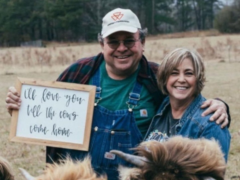Krista+Fuller+-+IMG_7739 Smiling couple posing outdoors on a farm, holding a sign that reads "I'll love you till the cows come home," with a scenic background of grass and trees. The man is wearing overalls and a cap, while the woman is in a denim jacket, both showcasing a warm, friendly atmosphere.